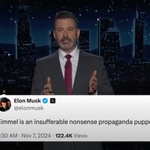 A man in a suit stands on a talk show stage. At the bottom of the screen is an X post from Elon Musk.